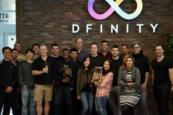 A group of around 17 people pose together in front of a brick wall featuring the "DFINITY" logo and infinity symbol, with two members holding small dogs. The image appears to be a team photo of DFINITY/ICP developers, suggesting the post is highlighting the irony that critics have gone silent following the UNDP × ICP partnership.