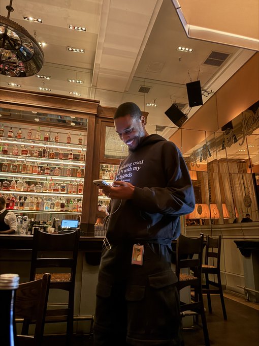 A young man in a dark hoodie smiles while looking at his phone inside an upscale bar, with bottles lining the shelves behind him and an ID badge clipped to his pants, reflecting the casual, long-form conversation about crypto the post describes.