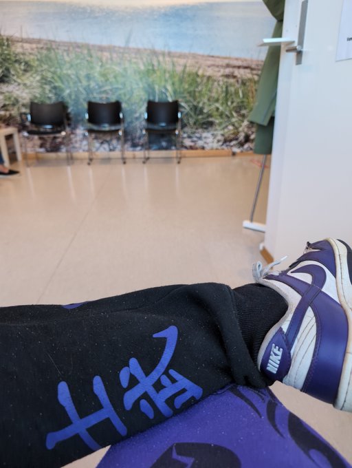 A first-person POV shot shows someone wearing blue and white Nike Dunk sneakers with black Nike sweatpants, appearing relaxed in what looks like a medical or office waiting area with black chairs against a nature-themed wall. The image complements the post's theme of awaiting a doctor's visit, with the giveaway serving as a distraction during recovery.