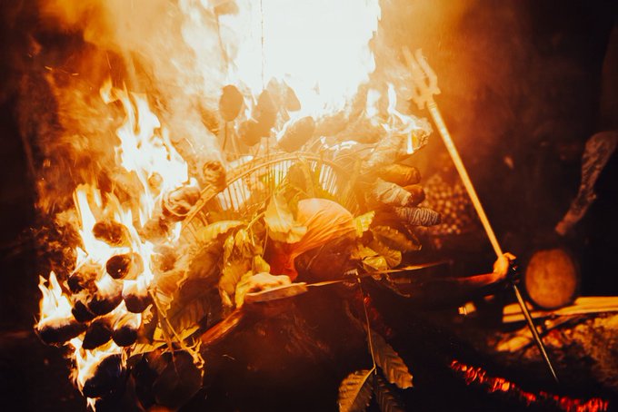 A dramatic close-up of a large bonfire at night, with intense orange flames engulfing wood, leaves, and what appears to be a metal frame or basket structure.
