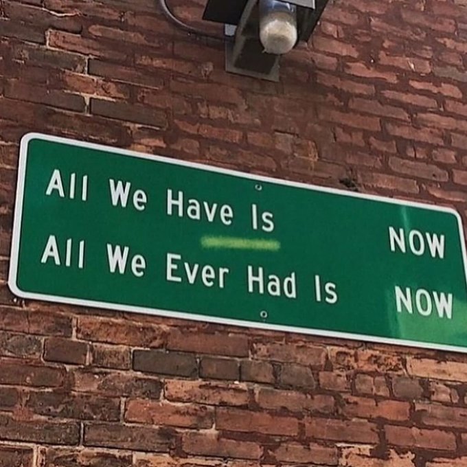 A green street sign mounted on a brick wall reads "All We Have Is NOW" on top and "All We Ever Had Is NOW" below, with a light fixture visible above it.