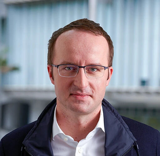 A man with short brown hair and glasses, wearing a dark jacket over a white shirt, stands in front of what appears to be an industrial or building background. The image relates to the accompanying text about Crypto.com CEO Kris Marszalek, who may be pictured here, addressing the security breach on the Tectonic protocol and the subsequent halting of the Cronos Network.