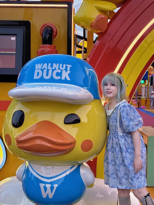 A young woman in a blue floral dress poses next to a large, smiling yellow duck mascot wearing a blue 'WALNUT DUCK' cap and jersey.