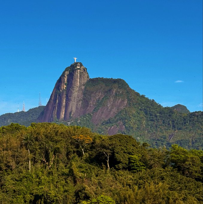 A photograph of a tall, rocky mountain with a statue of Christ on top, surrounded by lush green forest under a clear blue sky.