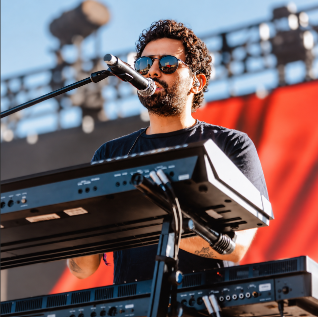 A bearded musician wearing reflective sunglasses and an in-ear monitor performs on stage, singing into a microphone while playing a keyboard and synthesizer setup under bright stage lights with a red backdrop. The image relates to the accompanying post about Jonathan Meléndez, the 38-year-old founding member and keyboardist of the Mexican band Camilo Séptimo, who was found dead in his family's apartment near Mexico City on September 1.