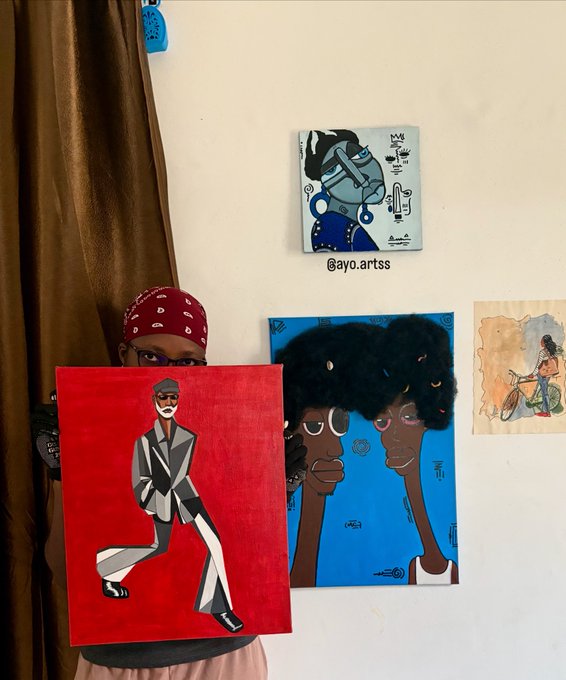 An artist wearing a red bandana holds up a vibrant red canvas featuring a stylized figure in a gray suit and sunglasses, showcasing their artwork in front of a wall displaying additional pieces—including a striking blue painting of two figures and the handle "@ayo.artss," perfectly matching their good morning greeting to fans.