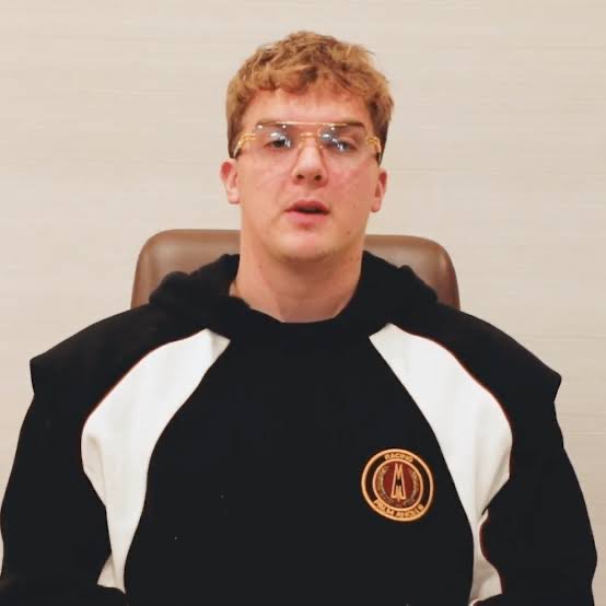 A curly-haired young man wearing clear-framed glasses and a black and white hoodie featuring an "A" logo sits in a brown chair against a light background, appearing to be speaking in what looks like a livestream or video recording. The image is presented in the context of a news announcement about Hunter Biden launching the $LAPTOP meme coin on Base.