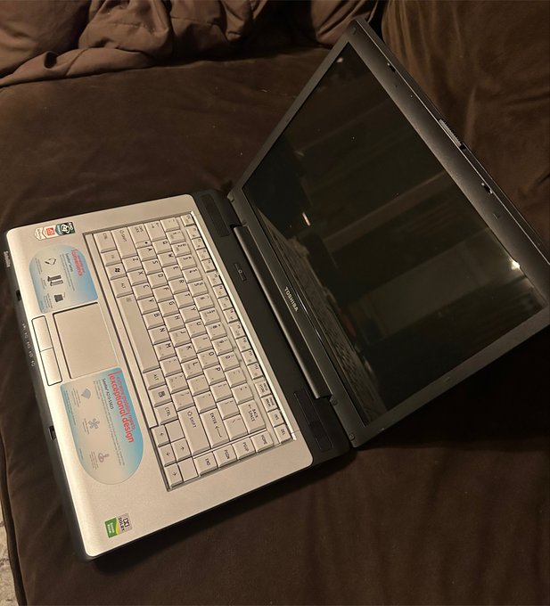 A photograph of an open Toshiba laptop resting on a brown bed, displaying Intel Core and NVIDIA stickers, with stickers on the palm rest and a dark, reflective screen.