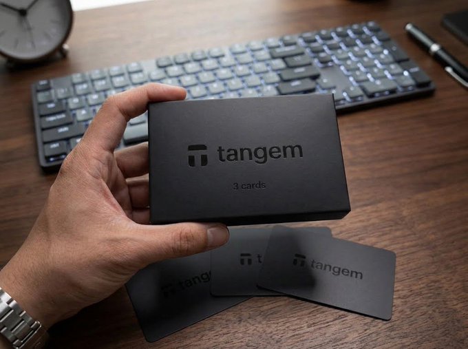 A hand holds a black box labeled "tangem 3 cards" over a wooden desk, with two matching black cards resting below it next to a backlit keyboard.