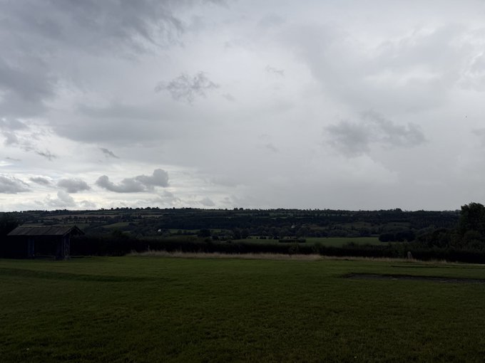 A wide landscape view shows a cloudy, overcast sky hanging over green rolling hills, fields, and a small wooden shed in the distance.