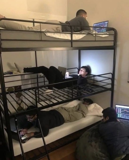 A dimly lit room with a metal bunk bed; multiple men are present, with one sitting on the top bunk using a laptop, one lying on the middle bunk, and another sitting on the bottom bunk.
