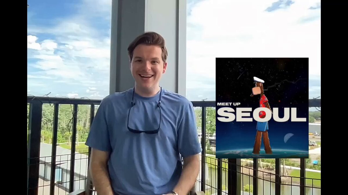 The image features a man wearing sunglasses standing next to a poster for Seoul. The man appears to be smiling at the camera while posing with the poster. The poster displays an illustration of a cartoon character, possibly from the movie "Moana". The background shows a cityscape or skyline, suggesting that the location might be in Seoul.
