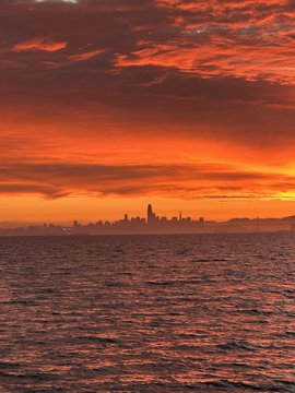 A dramatic sunset over a city skyline (with a distinctive tall tower) viewed across rippling water, with vivid red and orange clouds filling the sky.