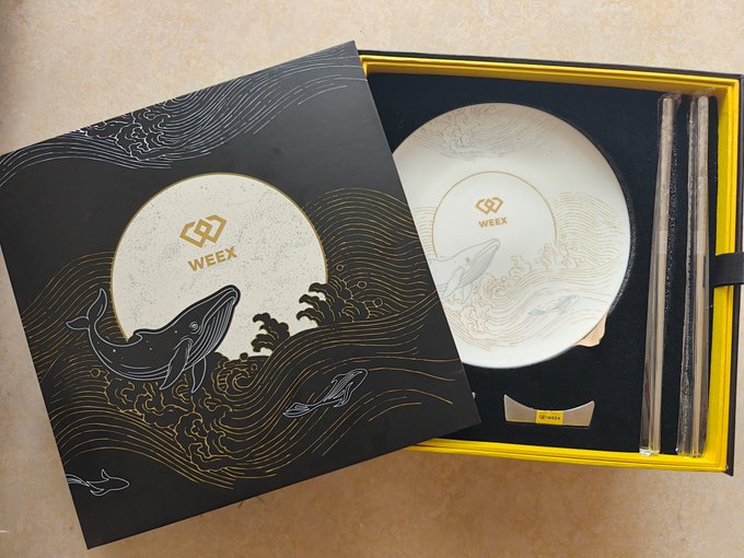 Photograph of a WEEX-branded gift set containing a gold-etched decorative plate featuring a whale design, presented in a black box with gold wave illustrations and accompanied by chopsticks.
