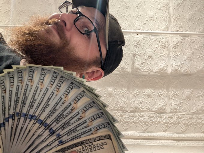 A bearded man wearing a cap and glasses poses with a large fan of hundred-dollar bills, looking upward against a decorative tiled wall.