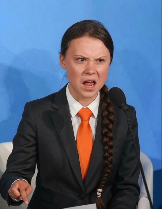 The image features a woman wearing an orange tie and black suit standing at a podium with her mouth open as if she's speaking or making a statement. The background of the image is blue, which contrasts with the woman's attire. There are no other objects in the scene besides the woman and the podium.