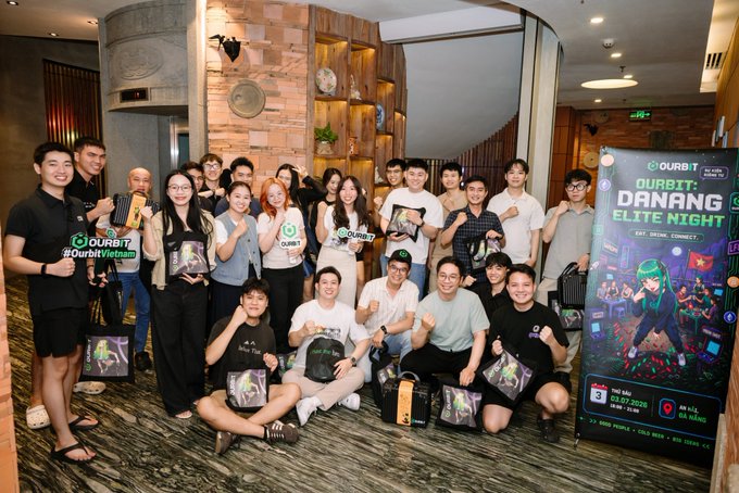 A group photo of attendees at the 'OURBIT DANANG ELITE NIGHT' event, posing with branded merchandise in front of a promotional banner featuring a character in green attire and event details including '03.07.2026'.