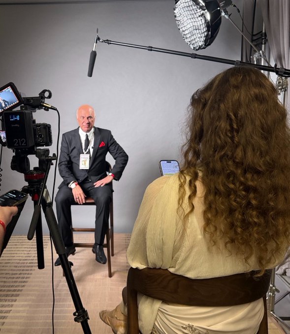 Behind-the-scenes photo of an interview setup showing a man in a gray suit with red pocket square seated against a gray backdrop, being filmed by a camera while a woman with long curly hair in a yellow top sits across from him holding a phone.