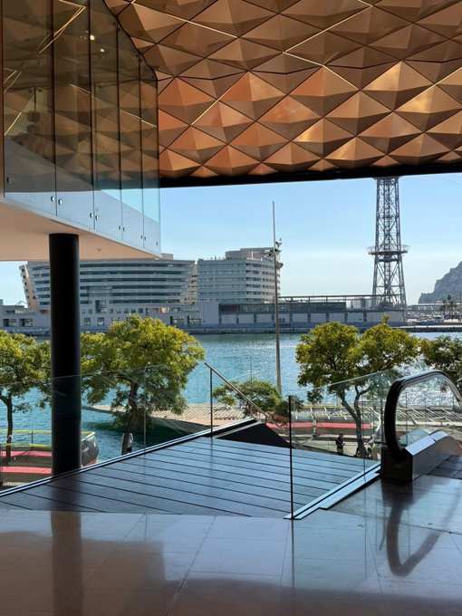 A view from inside a modern building with a geometric copper-colored ceiling, looking out through large windows toward a waterfront with a crane tower and buildings in the distance.