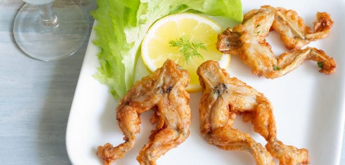 Plate of fried frog legs garnished with lettuce and a slice of lemon, sitting next to a wine glass.