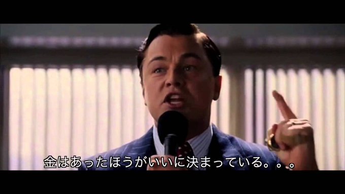 A cinematic still of Leonardo DiCaprio as Jordan Belfort from 'The Wolf of Wall Street' speaking at a microphone with Japanese subtitles reading 'It was decided that money is better.'