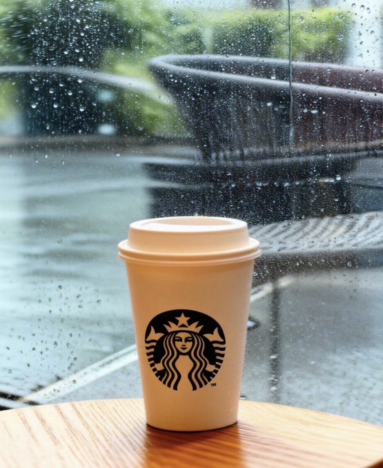 A beige Starbucks coffee cup with a lid sits on a wooden table, viewed through a window covered in rain droplets with dark outdoor chairs visible in the blurred background.