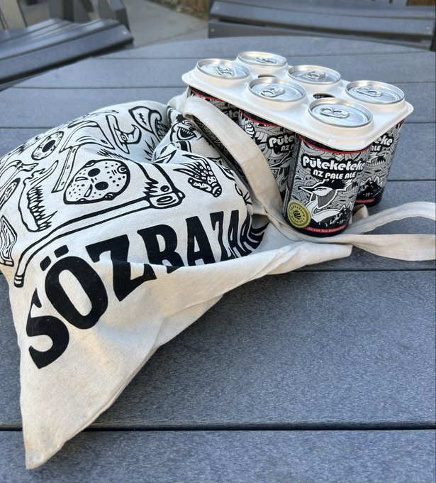 A tote bag displaying "SÖZBATAR" and a pack of "Püsketek" NZ Pale Ale sit on a wooden deck. The scene suggests a relaxed morning enjoyed with a craft beer, fitting a casual "good morning" vibe.