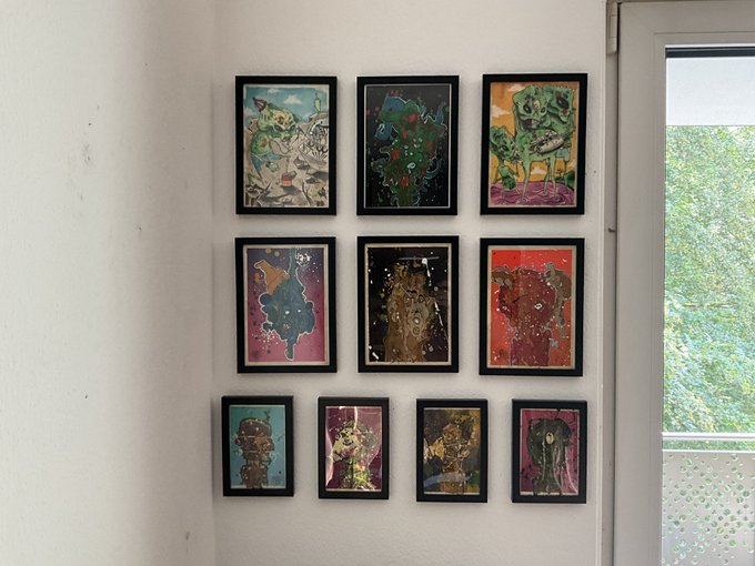 Ten framed abstract paintings are arranged on a white wall next to a window with trees visible outside.