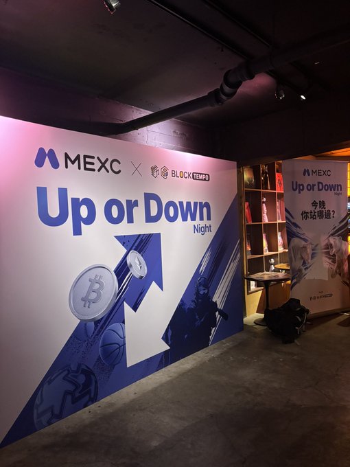 An event space with a large promotional backdrop for MEXC and BLOCKTEMPO featuring 'Up or Down Night' with an arrow graphic, alongside display shelves with merchandise.