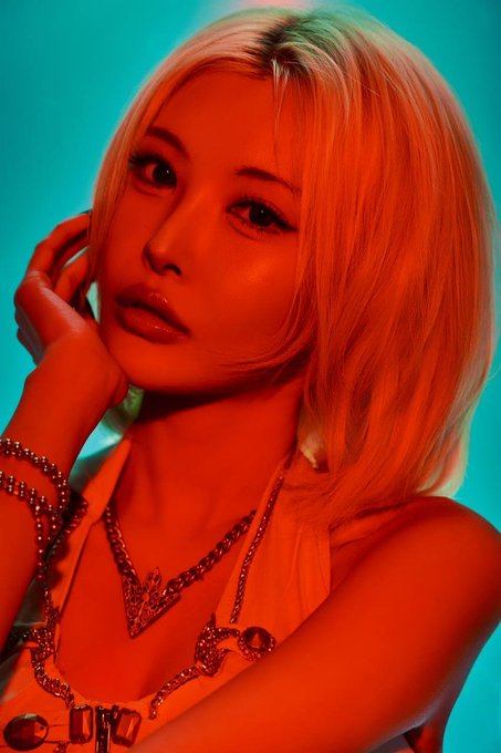 A stylized portrait features a young woman with short blonde hair bathed in intense red and orange lighting against a teal background, wearing layered silver chain necklaces.