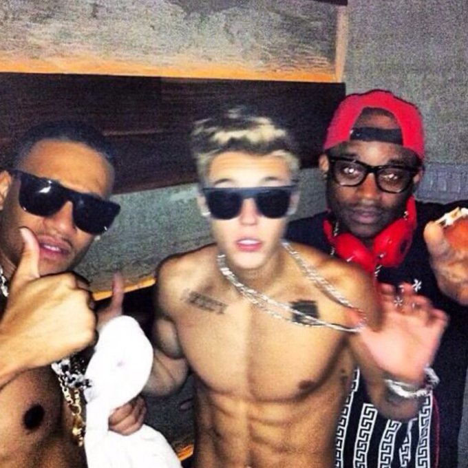 Three men, one shirtless wearing dark sunglasses and silver chains, pose together in what appears to be a recording studio.