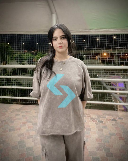 A young woman with long dark hair stands wearing a grey oversized t-shirt featuring a light blue abstract logo, photographed outdoors at night near a railing.