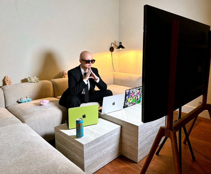 A bald man in a black suit and sunglasses sits on a beige couch with two laptops, watching a large screen, with a can of energy drink on the table.