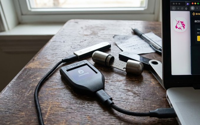A Trezor cryptocurrency hardware wallet sits on a wooden table connected to a laptop, with a USB drive, earbuds, scissors, a pen, and a handwritten note nearby.