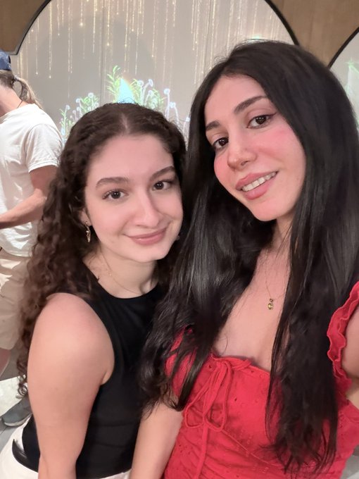 A selfie of two young women smiling together, one with curly brown hair in a black top and the other with long dark hair in a red ruffled top.