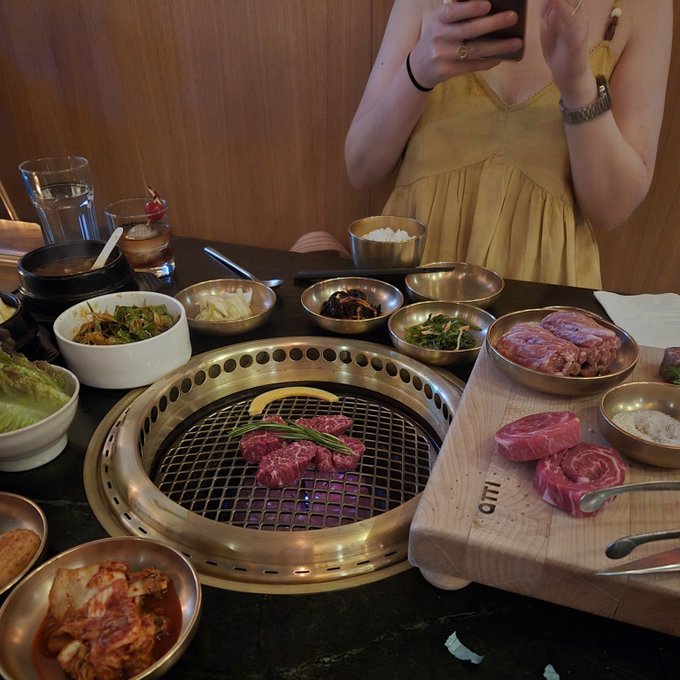 A person takes a photo at a Korean BBQ table spread with various banchan (side dishes) and raw meat being grilled on the built-in grill, with one cutting board labeled "ETON" visible — capturing a birthday feast for @CallMeChenAgain.