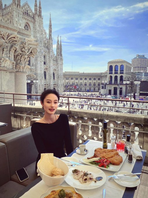 A young woman in a black top sits at an outdoor restaurant table with food and drinks, with the Milan Cathedral (Duomo di Milano) visible in the background.