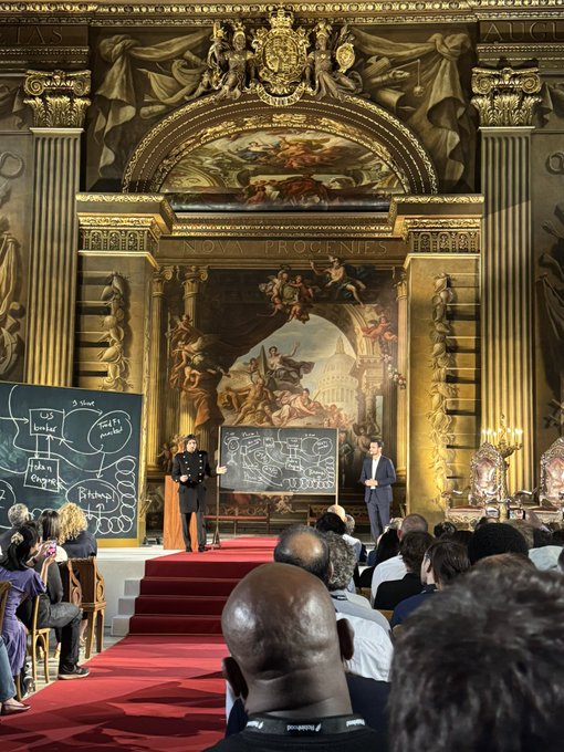 Two presenters stand on stage in an ornate, gilded hall featuring a large classical painting and the inscription "NOVA PROGENIES," presenting in front of chalkboards covered in handwritten diagrams while an audience watches from the red-carpeted floor. The grand setting and formal presentation align with the post's description of a major product launch event for Robinhood, Arbitrum, and Offchain, capturing the milestone moment of unveiling new offerings to the audience.
