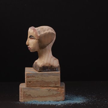 A small sculpted bust of a figure with an Egyptian-style headdress mounted on a tiered wooden-looking base, set against a dark background with blue powder scattered at the base.