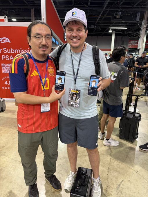 Two men posing for a photo at what appears to be a convention; the man on the left wears a red and yellow soccer jersey, while the man on the right wears a gray shirt and 'Always Forward' cap, both showing smartphone apps on their screens.
