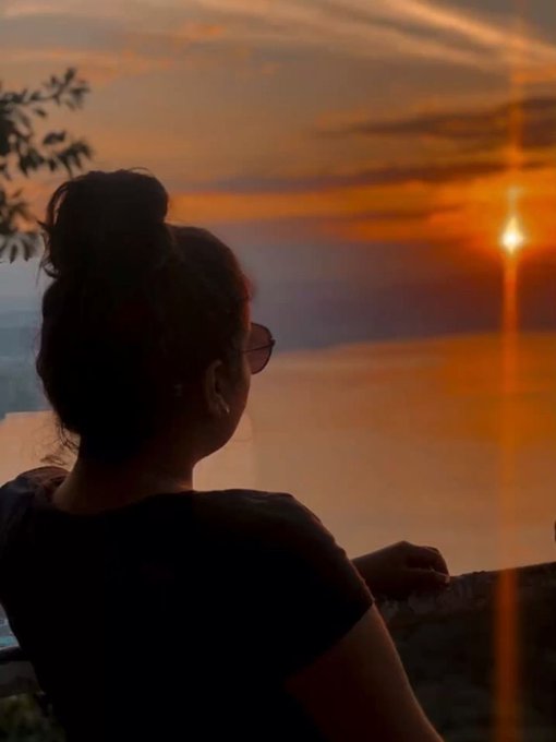 Silhouette of a woman with a bun and glasses watching a vibrant sunset, with the sun's reflection visible on water in the background.