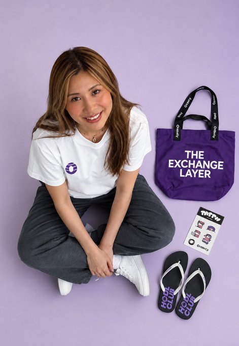 The image features a woman sitting cross-legged with her legs crossed, wearing a white t-shirt and black pants. She has a purple bag next to her that says "The Exchange Layer" on it. There are also two pairs of flip flops in the scene - one pair is purple and the other is white. The image includes text elements such as "Orderly 作为 Web3 Exchange Layer 太强了！ 共享统一 Order Book ，全链深度 CEX 级零代码 就能拥有自己的 Perp DEX ，流动性直接拊满，收入全归自己". The woman is smiling, and the purple bag has a "The Exchange Layer" design on it.