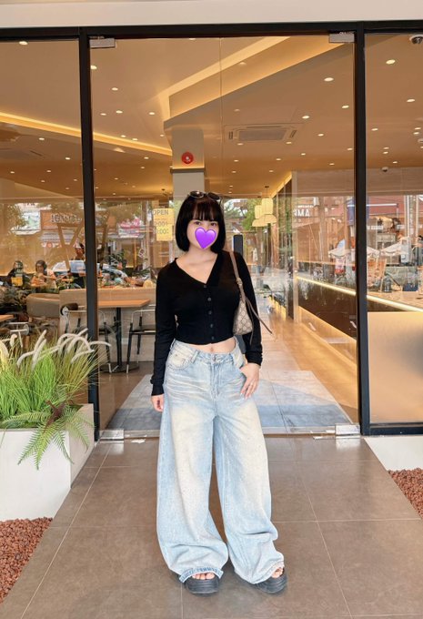 A young woman with dark hair and bangs stands outside a cafe, wearing a black long-sleeve cropped top and oversized light-wash blue jeans, with sunglasses resting on her head.