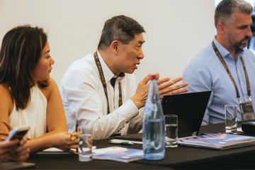The image shows a group of people sitting around a table, engaged in conversation or working together. There are three individuals visible - two men and one woman. One man appears to be speaking while holding a water bottle, possibly discussing an issue related to hydration during their meeting. The other two individuals have laptops on the table, suggesting that they might be working on a project or sharing information with each other. A cell phone is also present on the table, indicating that someone in the group may be using it for communication purposes.