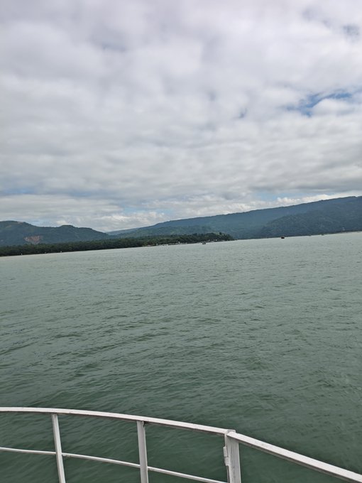 A view from a boat's white railing looking out over a calm, greenish body of water toward a distant shoreline of forested hills under a cloudy sky.