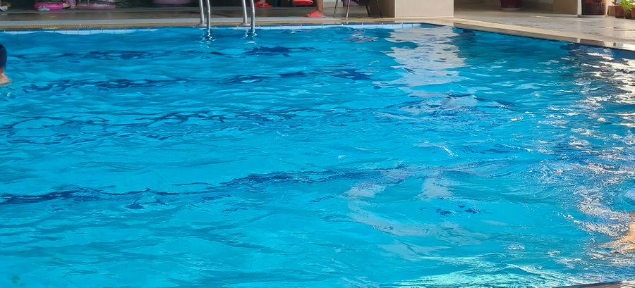Close-up view of bright blue swimming pool water with gentle ripples, showing pool ladder railings and a partial view of a person's arm on the left edge.