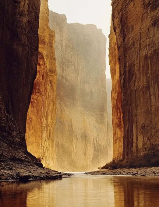 A scenic landscape of a narrow canyon with towering golden-brown rock walls and a calm river flowing through the bottom, lit by soft sunlight.
