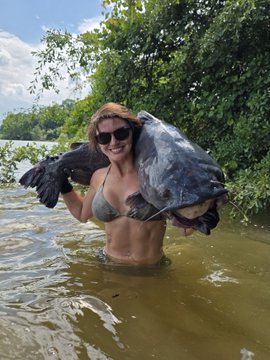 A woman in a bikini and sunglasses stands waist-deep in a river, holding up a large fish she has caught, smiling for the camera—fitting the "#GetBit" theme of celebrating a successful catch after a long absence from posting.