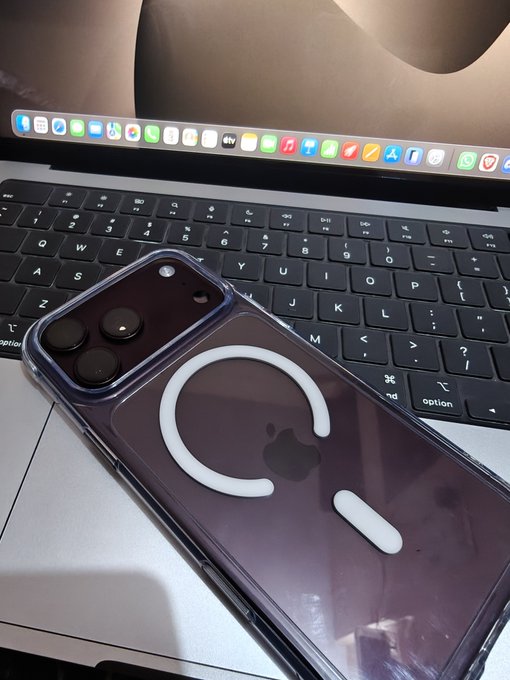 A clear iPhone case with MagSafe ring and white charging indicator rests on a MacBook keyboard, with the laptop screen and dock visible in the background.