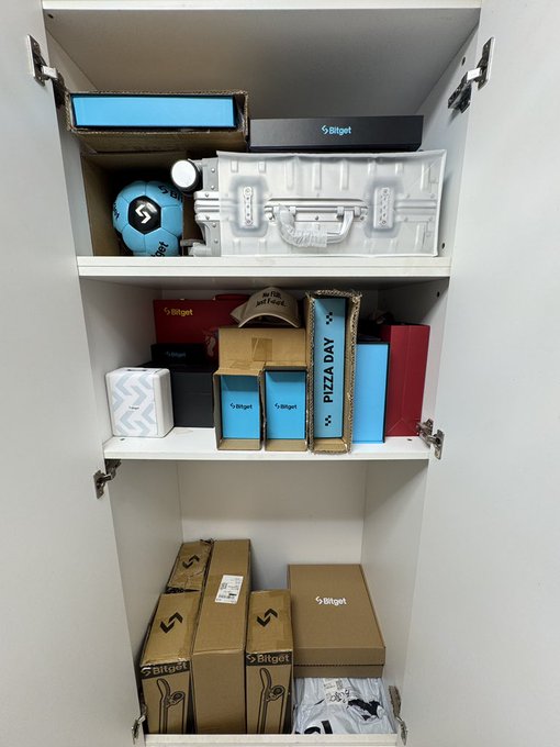 A white cabinet or shelving unit filled with various boxes and packages branded with the 'Bitget' logo, along with a silver metallic briefcase and a soccer ball on the top shelf.